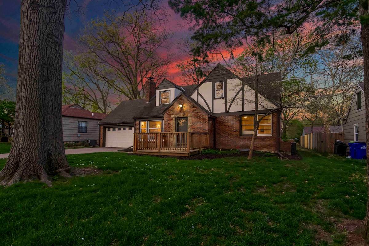 A charming two-story home featuring a mix of brick and wood exterior. A welcoming front porch with wooden railings is complemented by lush green grass, large trees, and a vibrant sunset sky. The driveway leads to an attached garage, enhancing the property's accessibility.