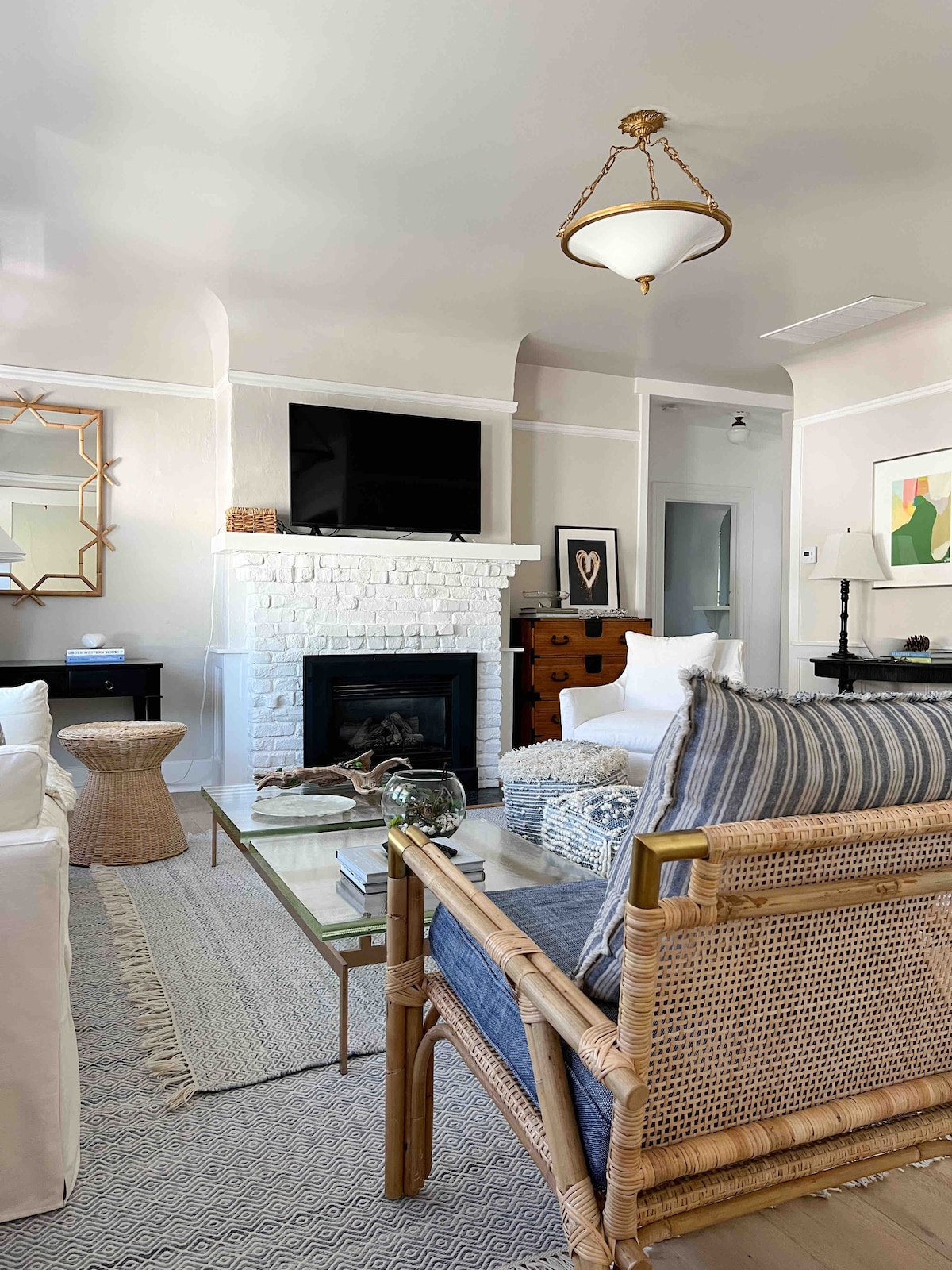 A light-filled living room features a white brick fireplace with a mounted television above it. Comfortable seating, including a rattan-accented chair and a white sofa, surrounds a glass coffee table. A textured area rug covers the hardwood floor, enhancing the cozy atmosphere.