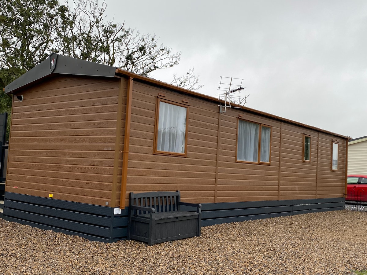 A lodge-style exterior is showcased, featuring wooden paneling and large windows. A bench is positioned in front, offering a place to sit. The structure is set on a gravel path, surrounded by trees, contributing to a rustic countryside feel.