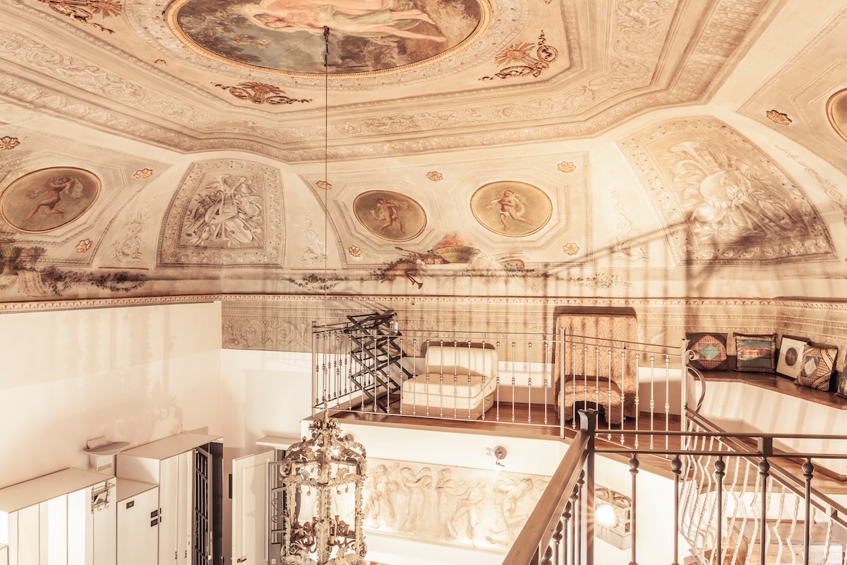 A high, frescoed ceiling displays intricate artwork and designs. The loft's elegant furniture can be seen, arranged thoughtfully. A wrought iron railing offers views of the open space below, where decorative elements contribute to the historical ambiance.