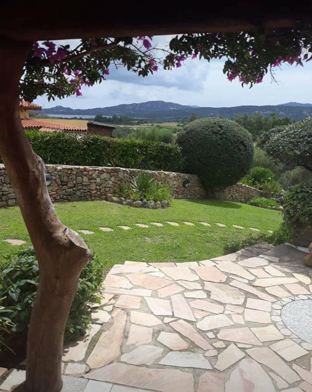 A garden layout features a well-manicured lawn bordered by flowering plants and stone pathways. Rolling hills are visible in the background, enhancing the serene outdoor setting. A stone patio with a unique design is situated in the foreground, framed by a wooden column and greenery.