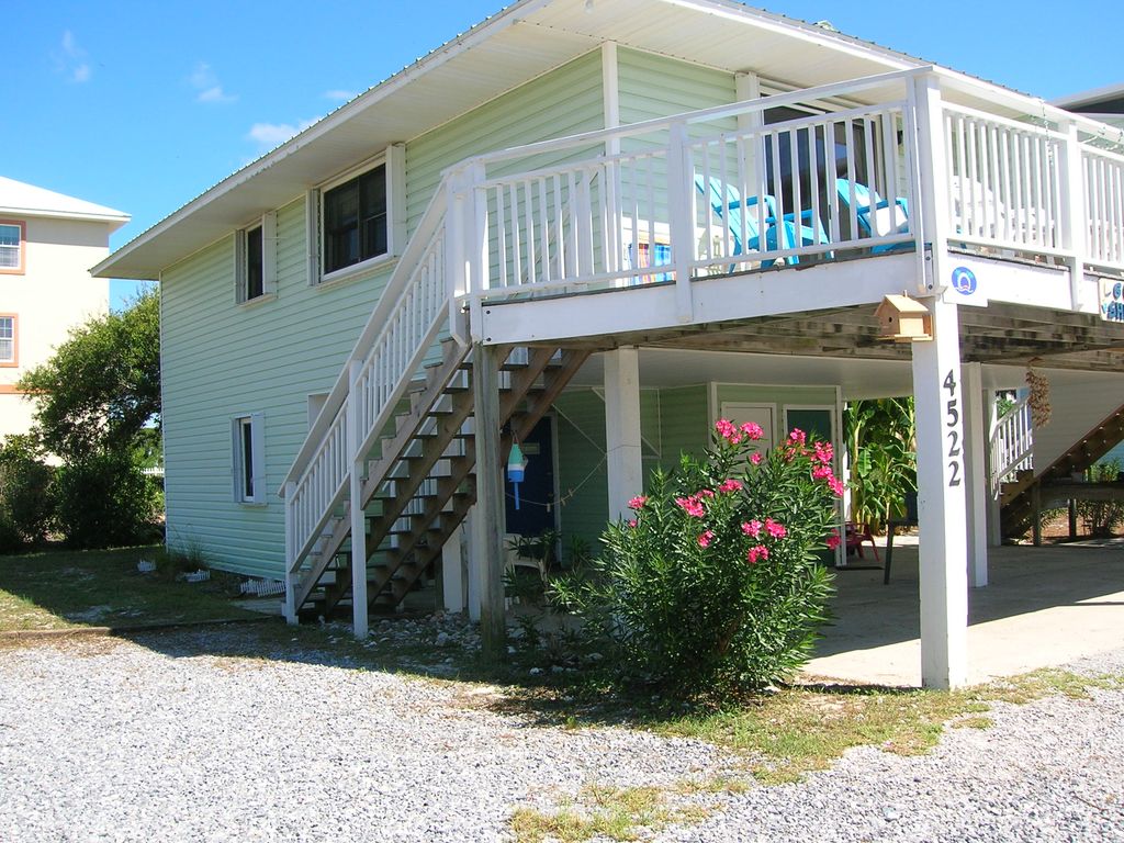 The exterior features a two-story beach house with light green siding and a covered balcony. A staircase provides access to the upper level, which is adorned with blue chairs. Lush greenery and pink flowers surround the base, creating a natural setting.