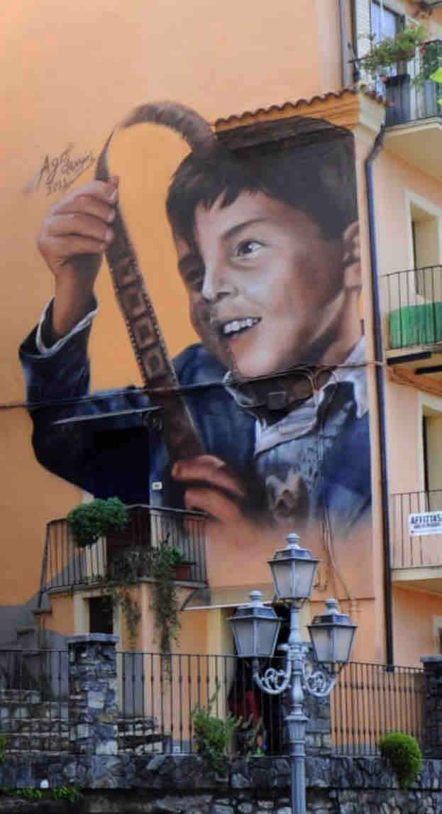 A large mural is depicted on the exterior of a building, showcasing a figure holding a curved instrument. The artwork spans multiple floors and is complemented by the surrounding architecture, including balconies and street lamps at the base.