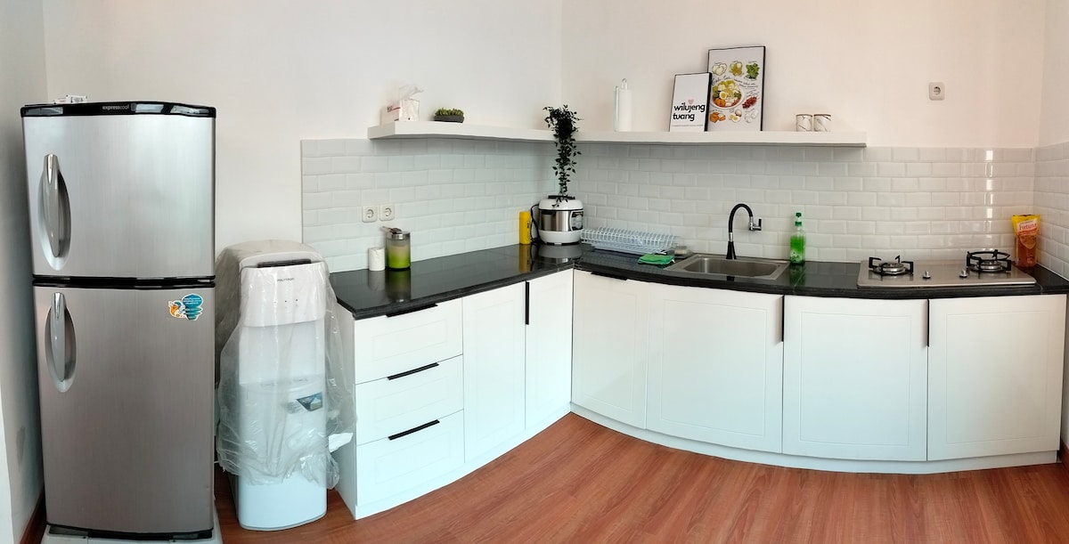 A modern kitchen space features sleek white cabinetry and a dark countertop. Stainless steel appliances, including a refrigerator and a gas stove, are visible. A water dispenser and various kitchen tools are neatly arranged, contributing to a functional cooking area.