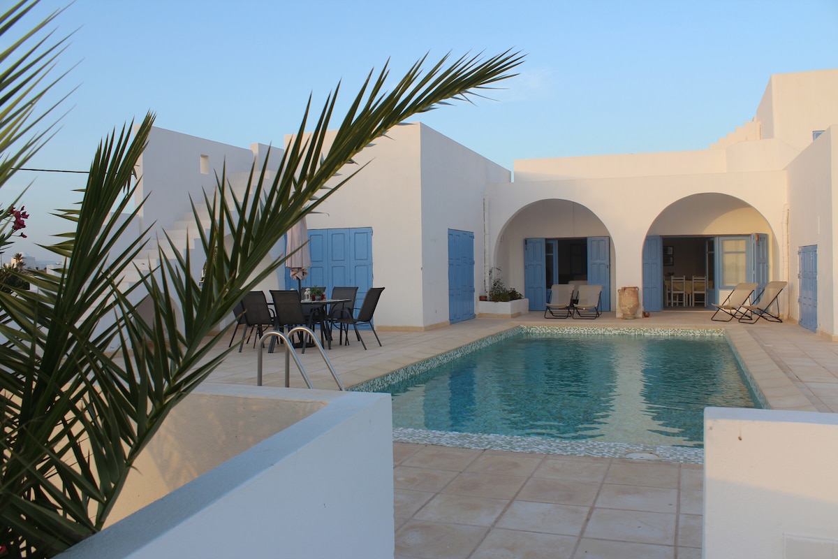 A clear pool is surrounded by a tiled terrace, with lounge chairs and a dining area. The whitewashed walls reflect the sunset, and arched doorways lead into the villa's interior. Lush greenery borders the space, contributing to the serene atmosphere.