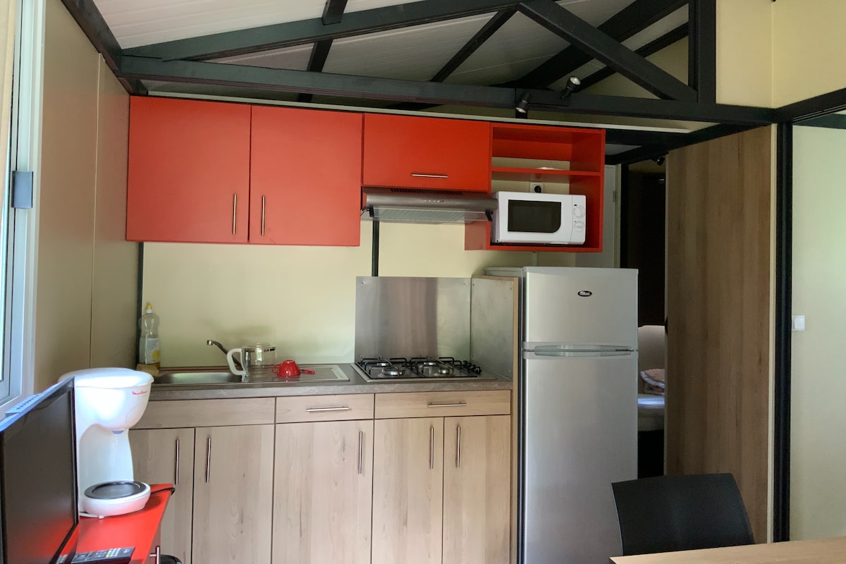 A compact kitchen area features red cabinetry and stainless steel appliances, including a gas stove, microwave, and refrigerator. A coffee maker sits on the counter, and a window allows natural light to brighten the space. An adjacent area with a dining table is partially visible.