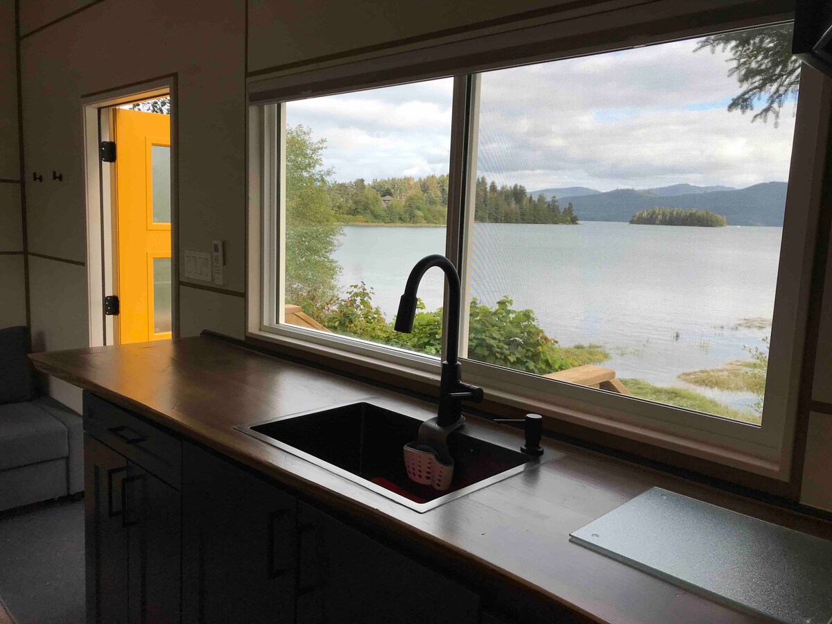 A modern kitchen area features a sleek black faucet above a stainless-steel sink, with natural wood countertops. A large window offers views of the serene waterfront, framed by greenery and distant islands, while soft lighting brightens the space.