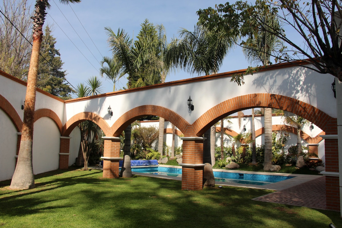 A serene outdoor area features lush green lawns, palm trees, and elegant arches. A swimming pool is visible, bordered by well-kept gardens. Soft natural light filters through the arches, enhancing the tranquil setting.