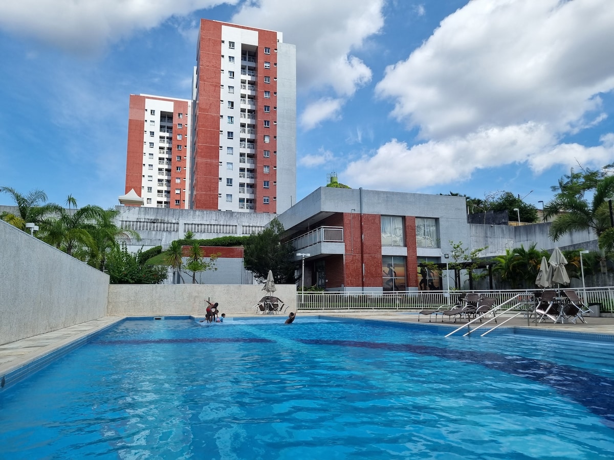 The image depicts a swimming pool surrounded by a stone deck. Several lounge chairs are arranged nearby. In the background, modern residential buildings rise against a partly cloudy blue sky, showcasing greenery around the pool area.