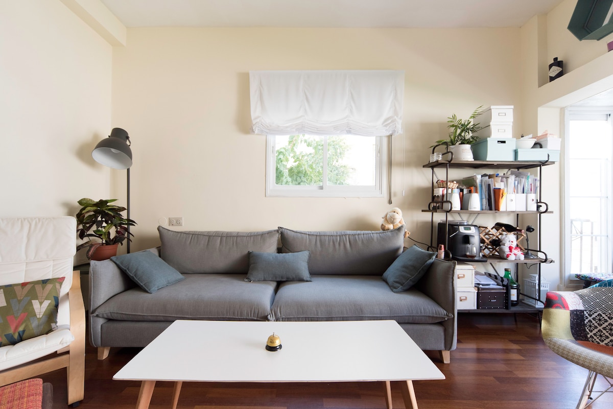 A spacious living area is captured, featuring a comfortable gray sofa adorned with accent pillows. A white coffee table is positioned in front, while natural light filters through the window above. Green plants and a shelving unit filled with various items add a touch of warmth to the space.