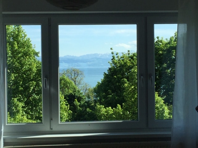 Three large windows offer a panoramic view of the serene Bodensee and the Swiss Alps in the distance, framed by lush green trees that enhance the scenery. Natural light fills the space, creating a bright and open atmosphere.