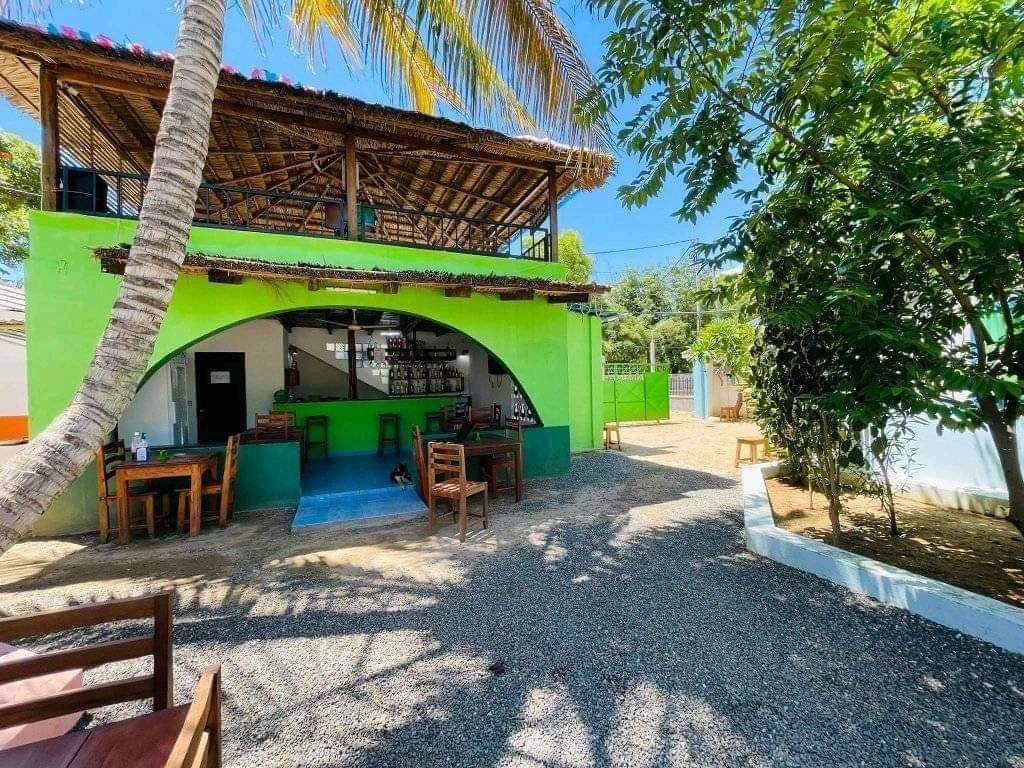 An outdoor dining area is set in a tropical environment, featuring wooden tables and chairs under a thatched roof. Surrounding greenery and a stone pathway create a serene outdoor setting, while a vibrant green structure serves as a backdrop.