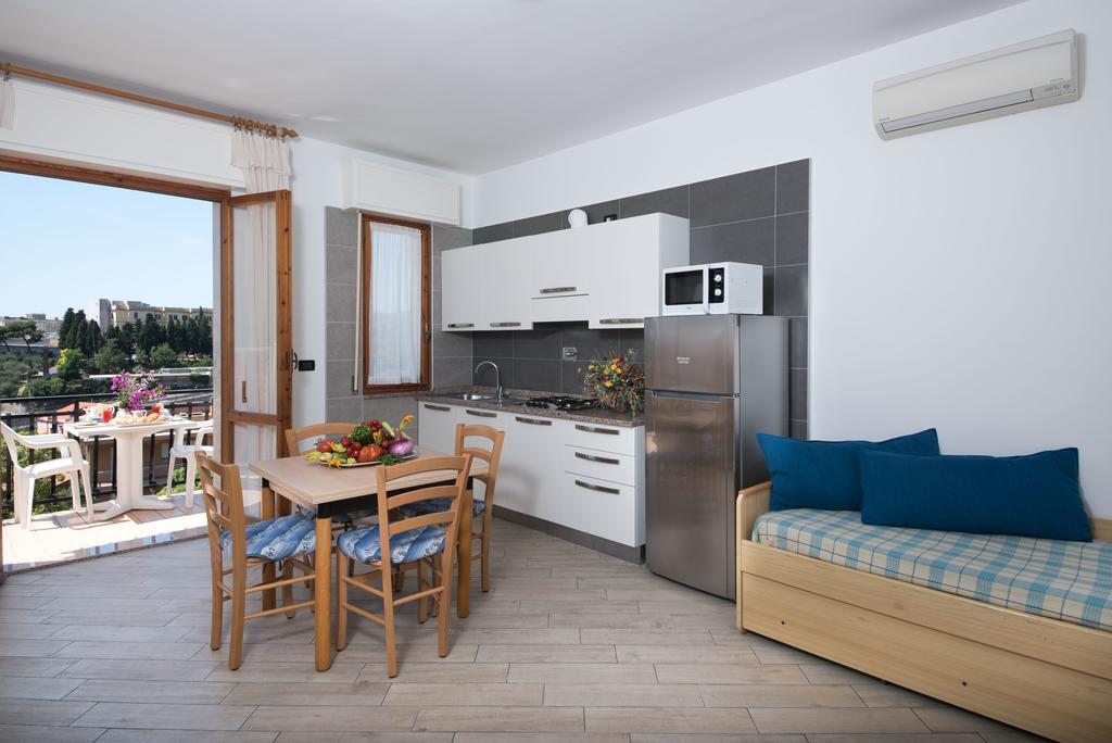 A bright living area features a sleek kitchenette with modern appliances, including a fridge and oven. A wooden dining table with four chairs is set beside a comfortable sofa bed. Natural light floods in through the open balcony doors, enhancing the welcoming ambiance.