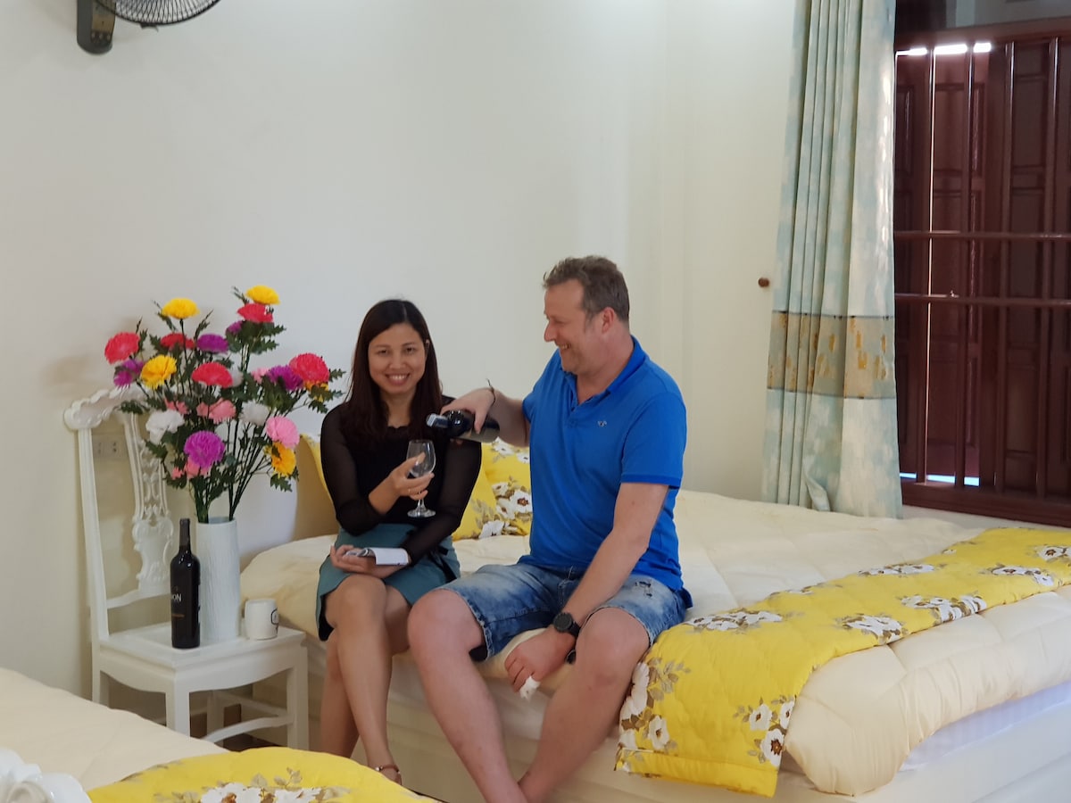 A spacious room is furnished with two beds draped in yellow bedding. A white chair accompanies a small table, adorned with a vibrant floral arrangement. Natural light enters through the window, highlighting the peaceful atmosphere.