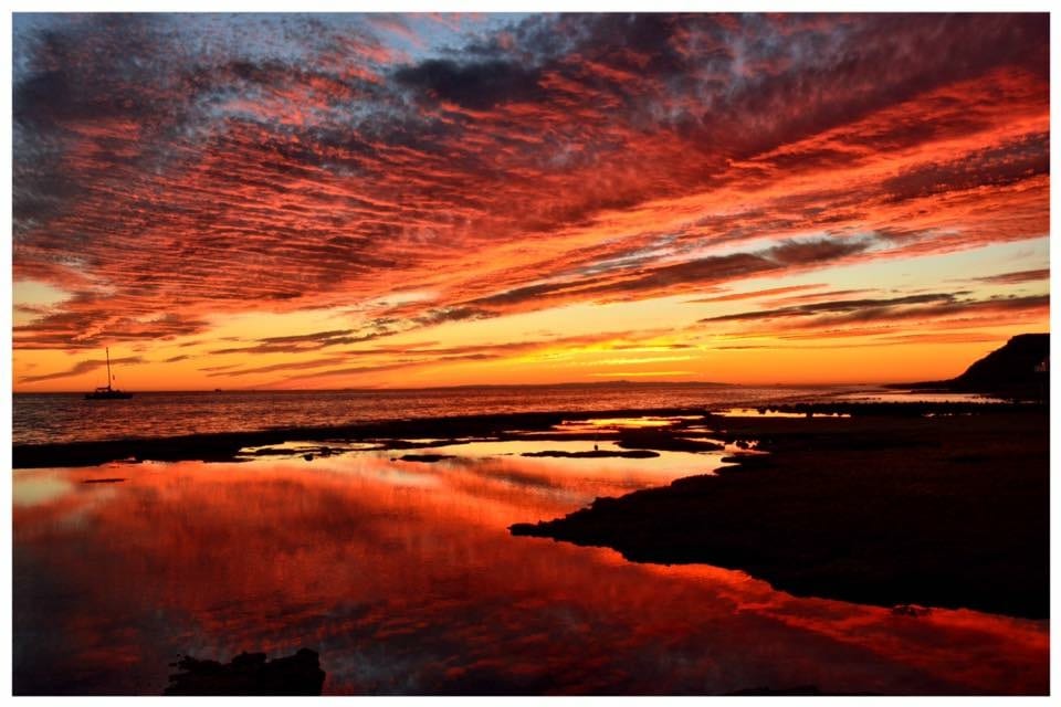 A vibrant sunset illuminates the sky with hues of orange, red, and purple, reflecting upon calm waters. The landscape features jagged rocks along the shoreline, emphasizing the tranquility of the seascape. A distant sailboat is visible against the backdrop.