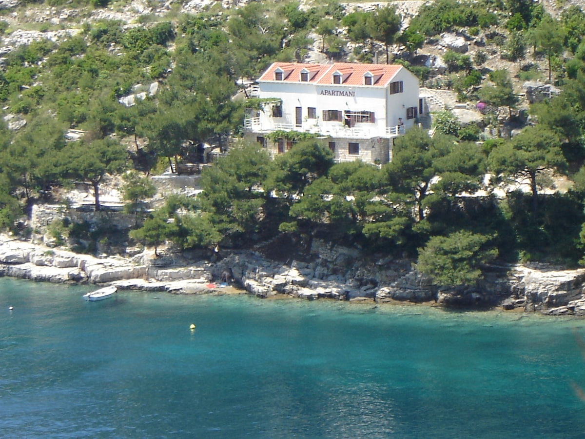A coastal building with a white exterior is nestled amongst trees, overlooking clear turquoise waters. The structure features multiple balconies and a red-tiled roof, with rocky shores visible at the base. The natural landscape provides a serene backdrop, enhancing the appeal of the location.