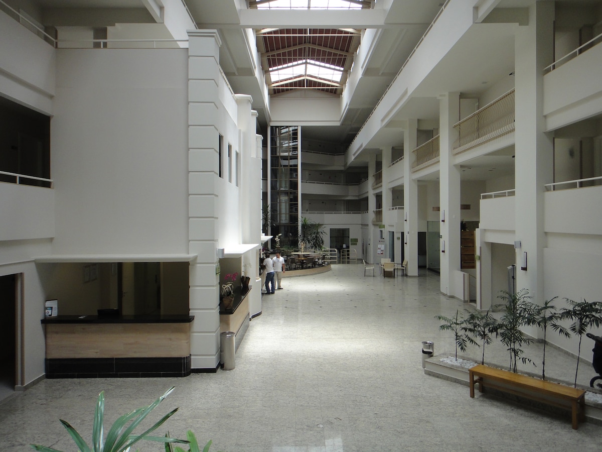 The spacious atrium features high ceilings and large windows, allowing natural light to fill the area. A reception area is visible to one side with a counter, while plants are located along the marble flooring, enhancing the open atmosphere.