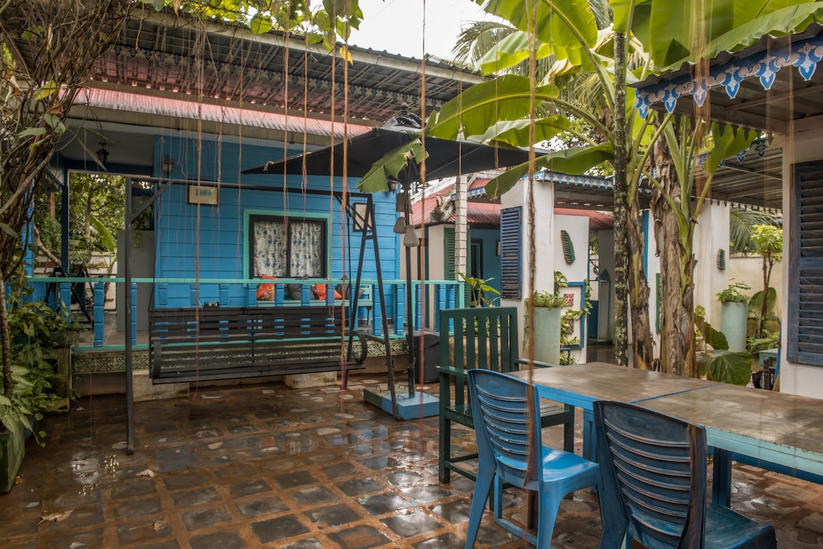 A tranquil outdoor space features a swing set and a wooden table accompanied by stacking blue chairs. Lush greenery and banana trees provide shade, creating a serene environment. The blue exterior of nearby buildings complements the natural surroundings.