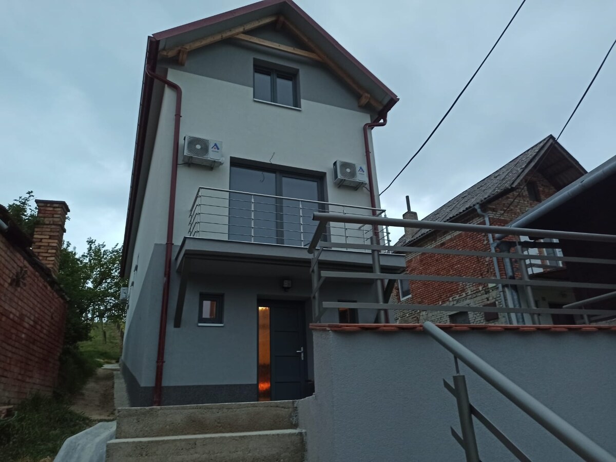 The modern two-story building is set against an evening sky, featuring a light-colored exterior and a covered entryway. A railing surrounds the outdoor staircase leading to the main entrance, which is highlighted by ambient lighting. Air conditioning units are visible on the façade.
