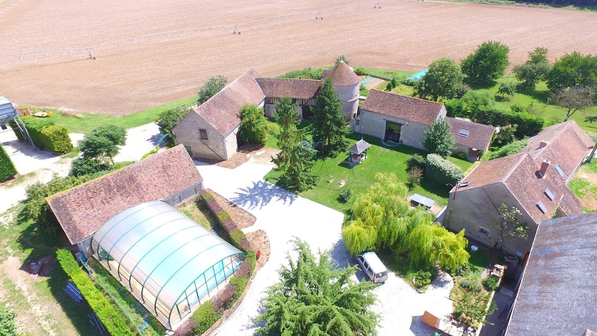 An aerial view showcases the expansive property featuring lush greenery and several well-maintained buildings. A covered pool area is visible, along with manicured gardens and open fields surrounding the estate, creating a serene and spacious environment.