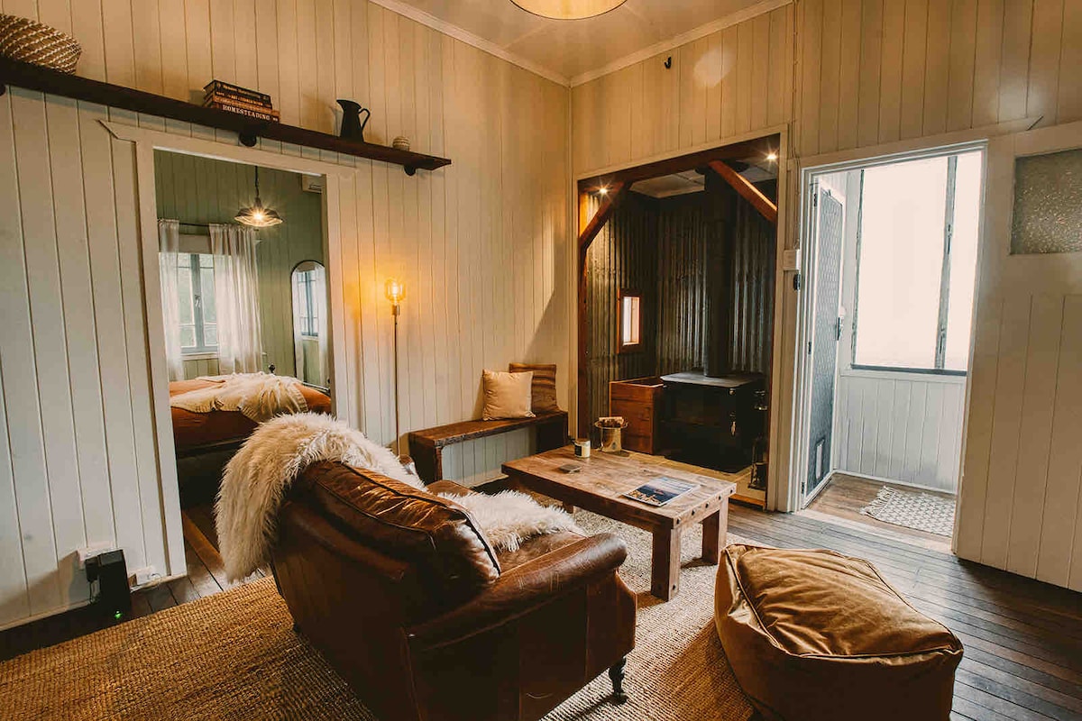 A comfortable living area showcases a plush leather sofa and a wooden coffee table, with soft lighting provided by a lamp. The adjacent room features rustic decor, and large windows offer views of the outdoors, enhancing the cozy ambiance of the space.