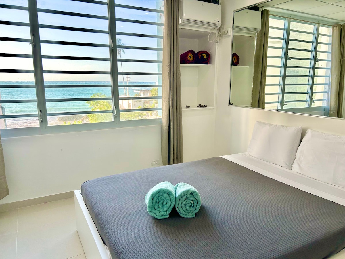 A bright bedroom features large windows with views of the ocean and palm trees. The spacious bed is neatly made with white linens, while two rolled towels in pastel blue are placed decoratively at the foot of the bed. Mirrored surfaces reflect natural light.