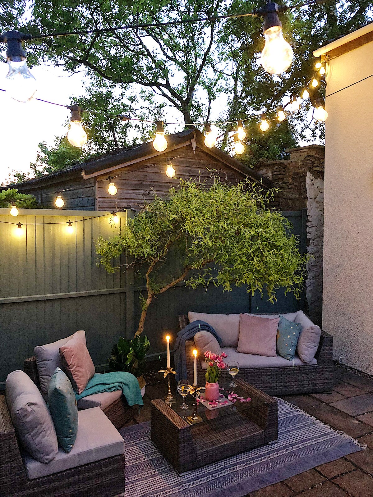 An outdoor seating area features soft cushions on a pair of contemporary sofas, surrounded by a low table. Warm string lights illuminate the space, creating a cozy atmosphere. A small tree and decorative plants add greenery against a backdrop of a wooden fence.