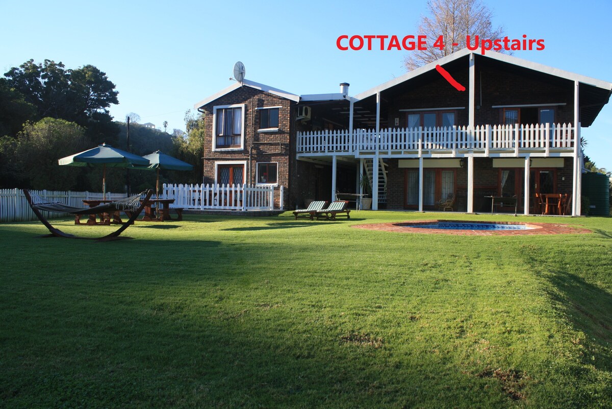 The exterior of a two-story cottage is displayed, featuring a brick façade and a balcony. A well-maintained lawn surrounds the property, with a hammock and outdoor seating visible. A communal swimming pool is nearby, enhancing the outdoor space. Clear skies and greenery complete the view.