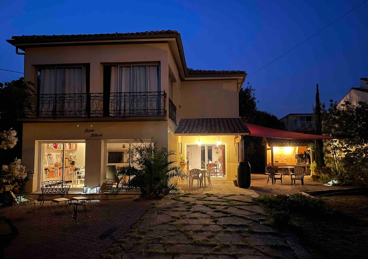 The two-story house is illuminated in the evening with warm light visible from the windows. Outdoor seating is arranged under a covered area, with plants surrounding the entrance. A path is seen leading to the house, contributing to the inviting ambiance.
