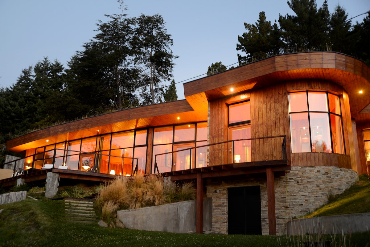 The modern house is showcased with large glass windows, reflecting warm light at dusk. The structure features a curved wooden facade and is surrounded by lush greenery, creating a harmonious connection with the landscape.