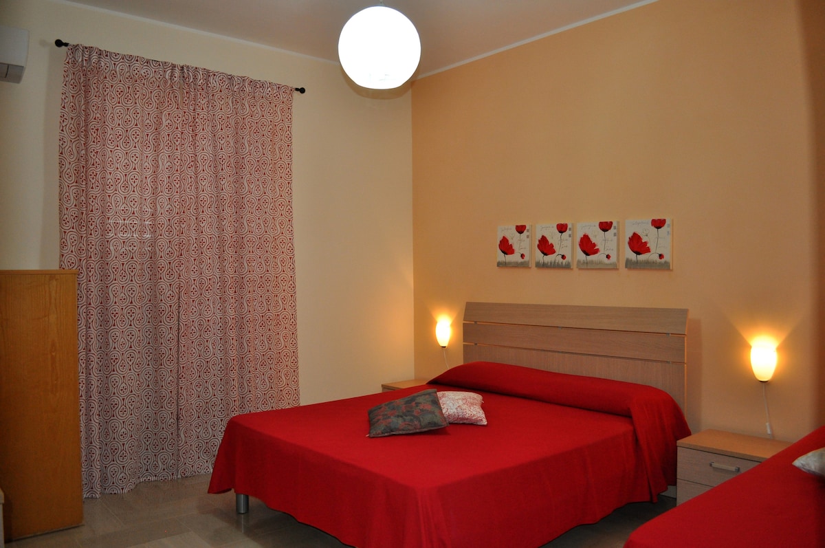 A warm-toned bedroom features two spacious beds draped in red covers. The walls are illuminated by soft light, complemented by a patterned curtain and decorative art pieces. A bedside table is located next to each bed, enhancing the room's functional layout.