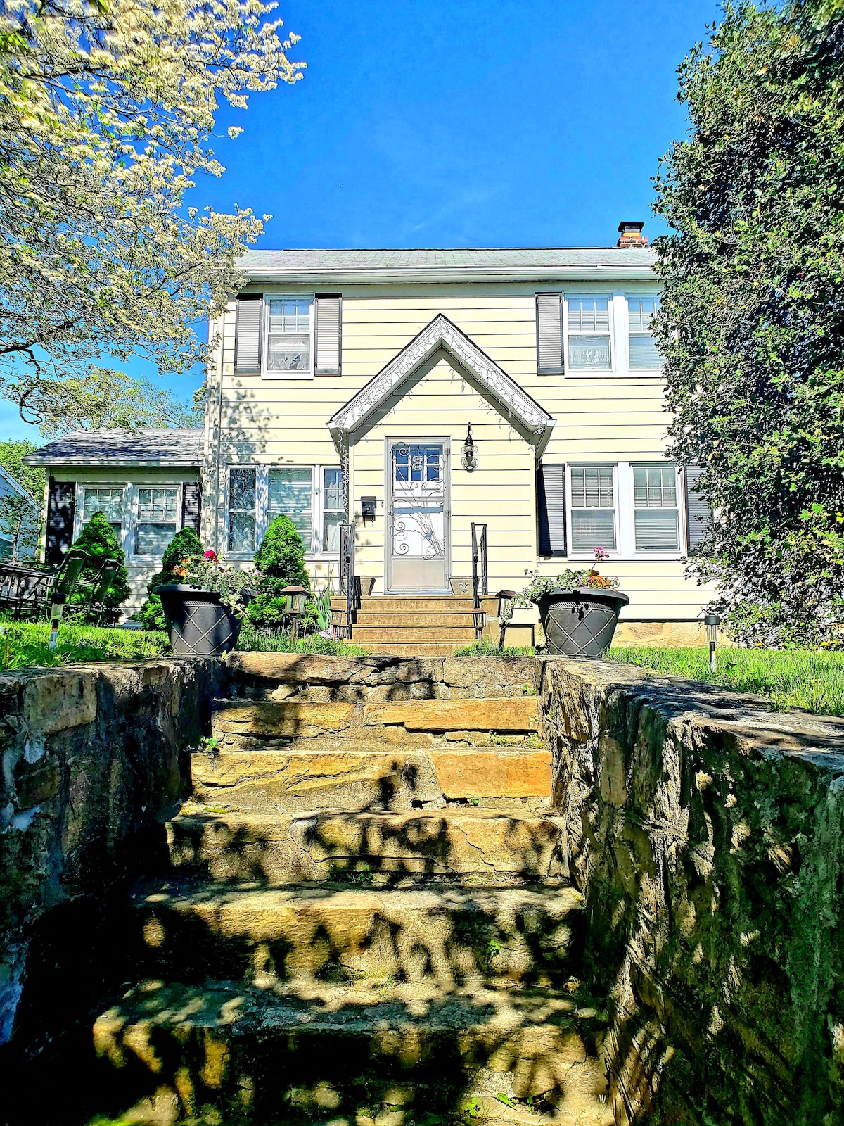 A charming yellow house is presented, featuring a symmetrical design with a peaked roof and prominent front door. Stone steps lead up to the entrance, framed by greenery and potted plants on either side. Clear blue skies create a fresh, inviting backdrop.
