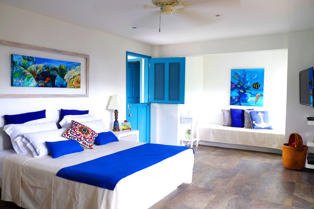 A spacious bedroom features a large bed dressed in crisp white linens with blue accents. A comfortable sitting area includes a light-colored sofa, complemented by vibrant wall art depicting sea life. Double doors offer access to natural light, creating an inviting environment.
