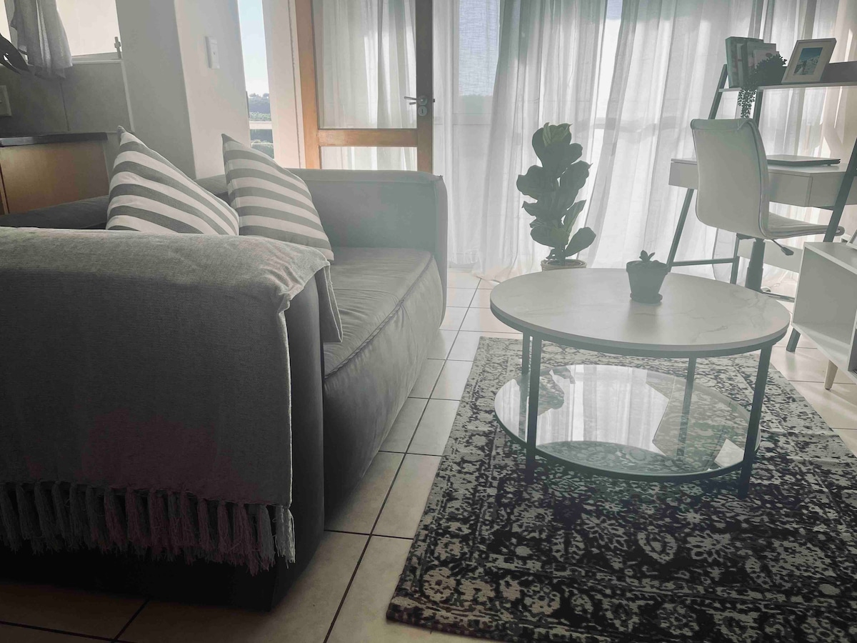 A light-filled living area features a grey couch with striped cushions and a round coffee table atop a patterned rug. Natural light filters through sheer curtains, illuminating a small plant on the table and a workspace with a chair and shelves in the background.