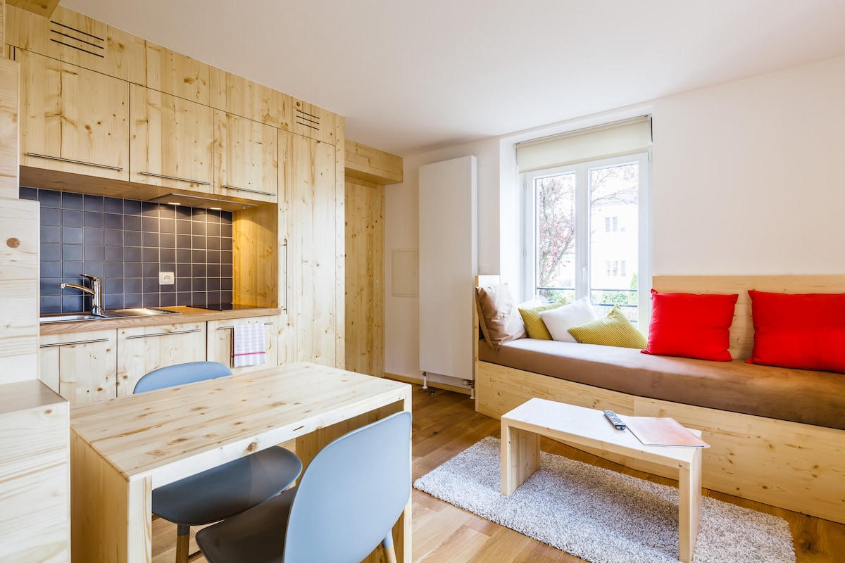 A contemporary duplex apartment features wooden cabinetry and an open kitchen area. A cozy seating nook with colorful cushions is positioned next to a window. A small dining table with four chairs is set against the warm-toned wood flooring, creating an inviting living space.