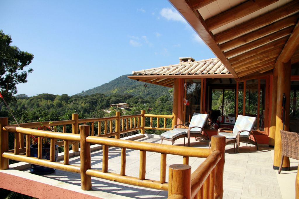 An expansive balcony offers views of the surrounding lush landscape. Comfortable lounge chairs are positioned to enjoy the scenery, complemented by wooden railings. The roof features wooden beams, enhancing the natural aesthetic of the space.