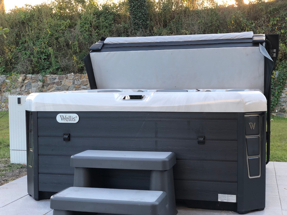 A spacious outdoor hot tub is positioned on a tiled patio, featuring a protective cover and steps for easy access. Lush greenery creates a calm backdrop, enhancing the tranquil setting. The hot tub is designed for relaxation and well-being.