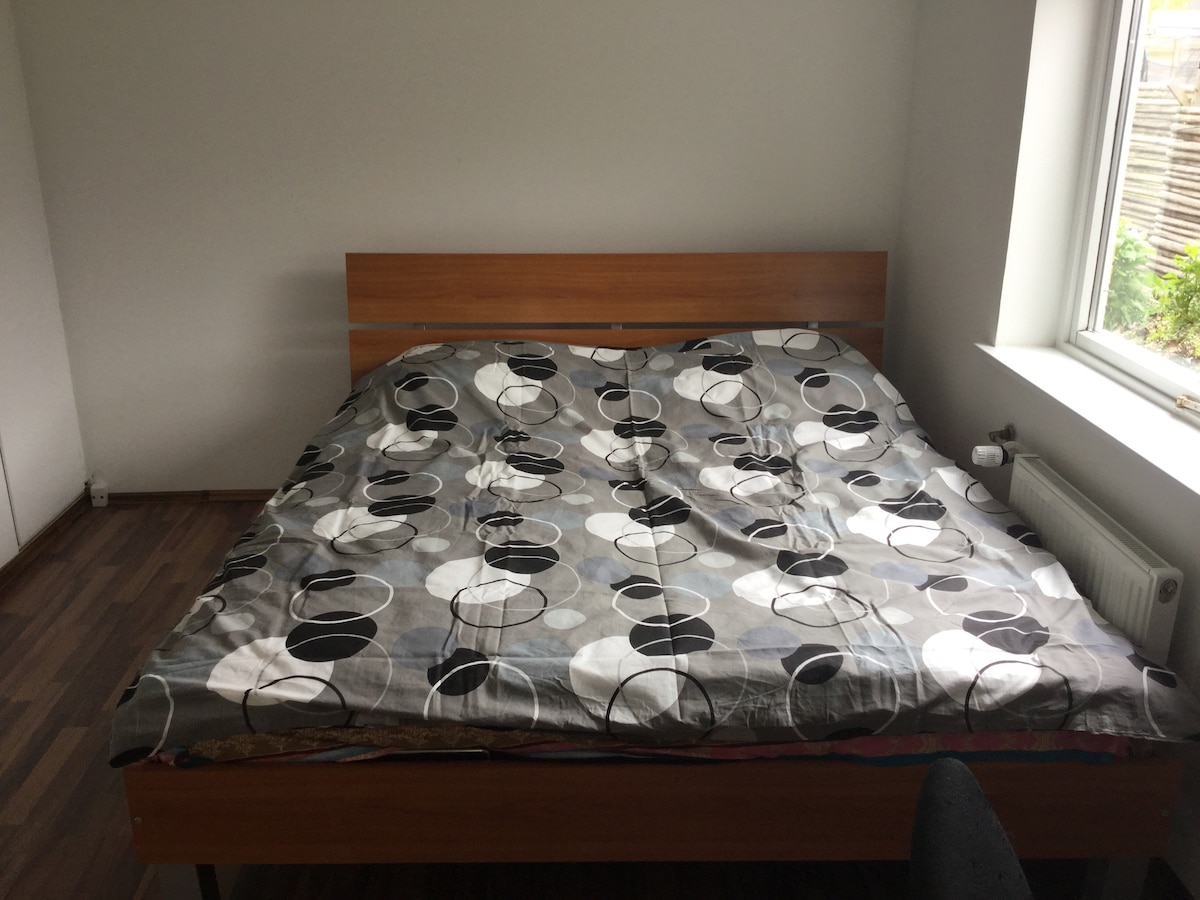 A spacious bed is positioned against a light wall, featuring a patterned comforter in muted colors. Natural light enters from a nearby window, illuminating the wooden floor and the simple design of the room.