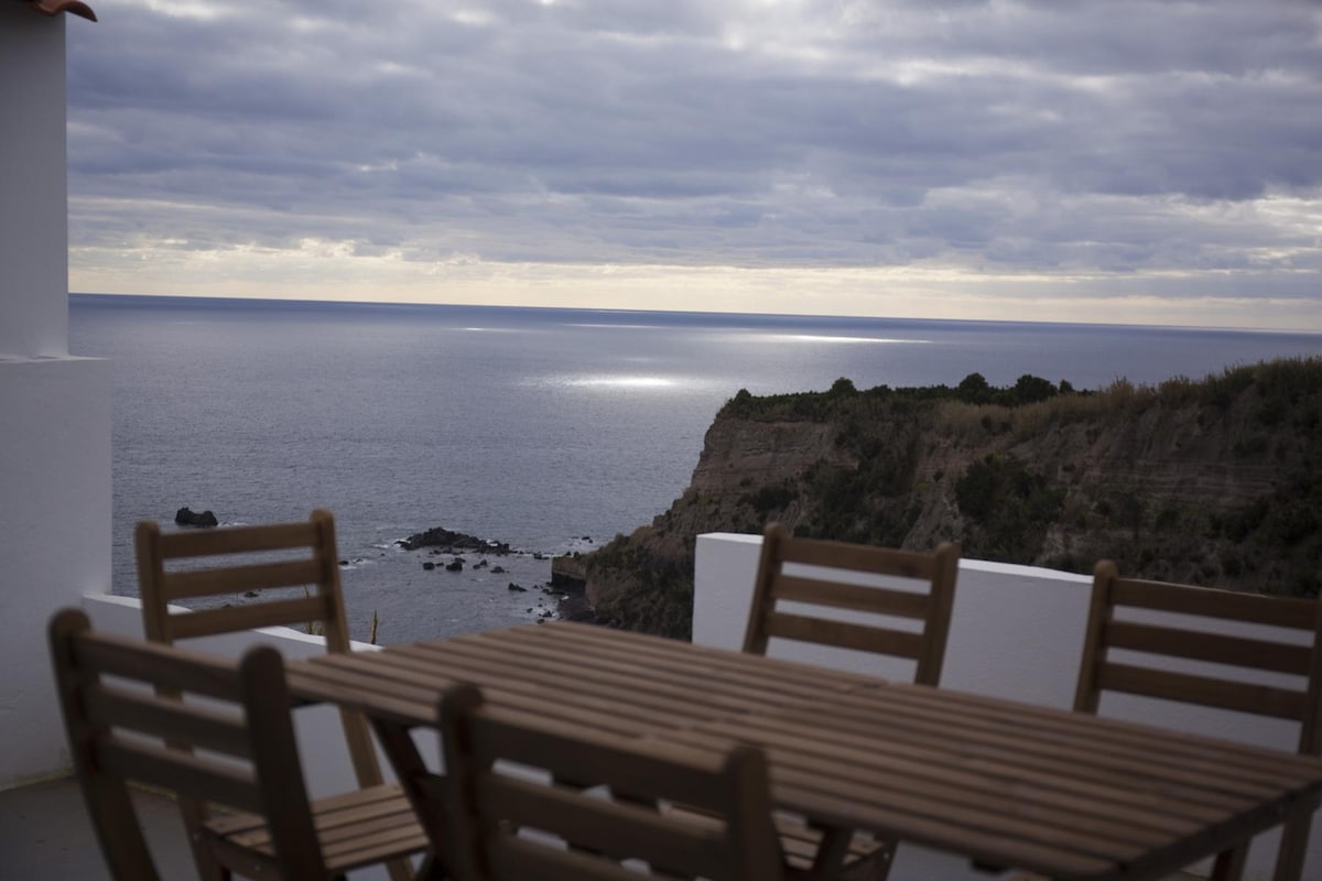 An outdoor terrace is equipped with a wooden dining table surrounded by multiple chairs. The scene overlooks a cliffside with views of the ocean, where the horizon meets cloudy skies. The tranquil waters reflect light, enhancing the peaceful coastal atmosphere.