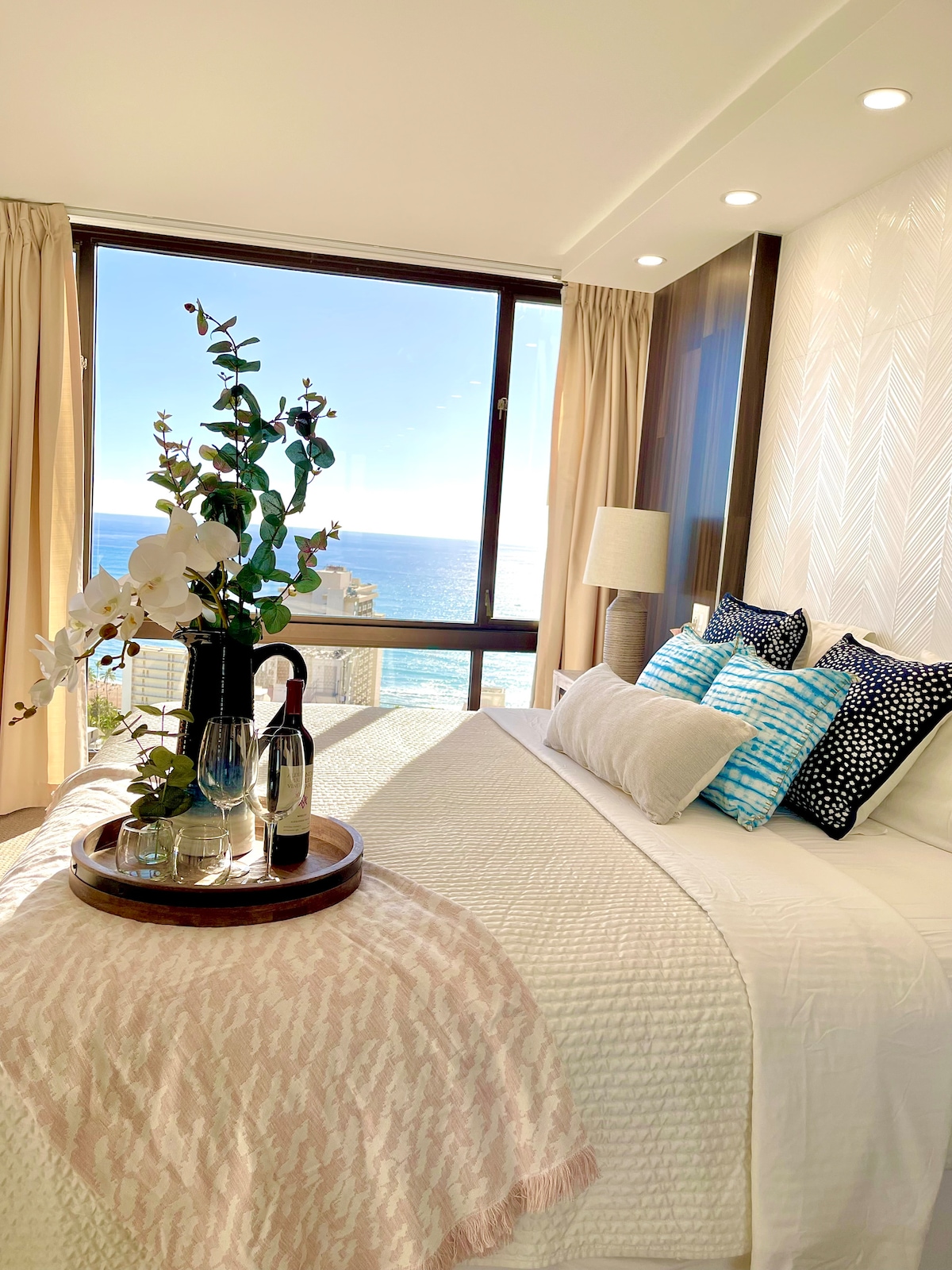A well-appointed bedroom features a spacious bed topped with decorative pillows in blue and white patterns. A tray with glasses and a bottle is displayed at the foot of the bed. Large windows offer a bright view of the ocean and natural light.