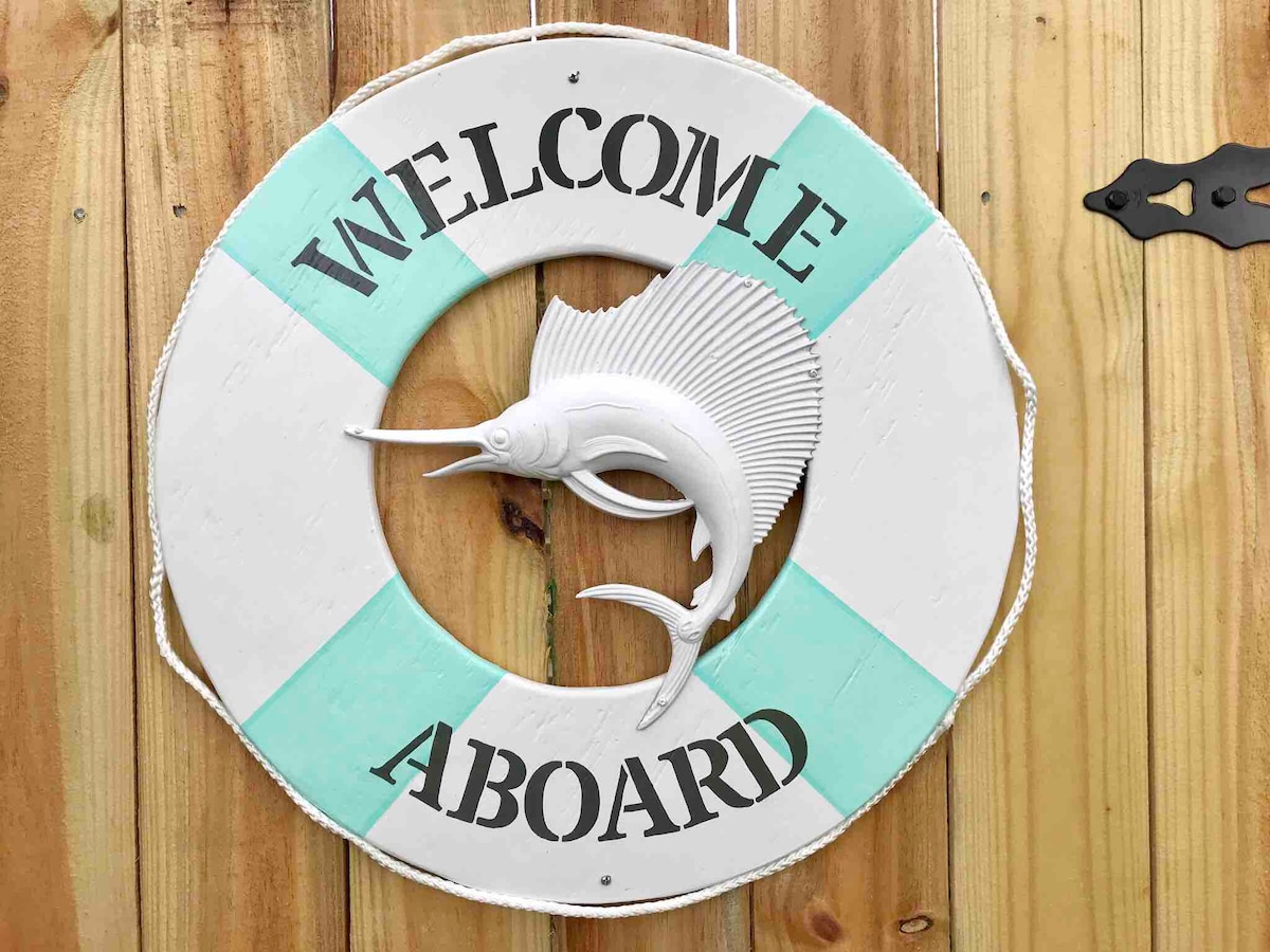 A decorative life preserver hangs on a wooden wall, featuring the words 'Welcome Aboard' in bold black letters. A stylized fish is displayed in the center, surrounded by mint green and white accents, inviting guests to a nautical-themed atmosphere.
