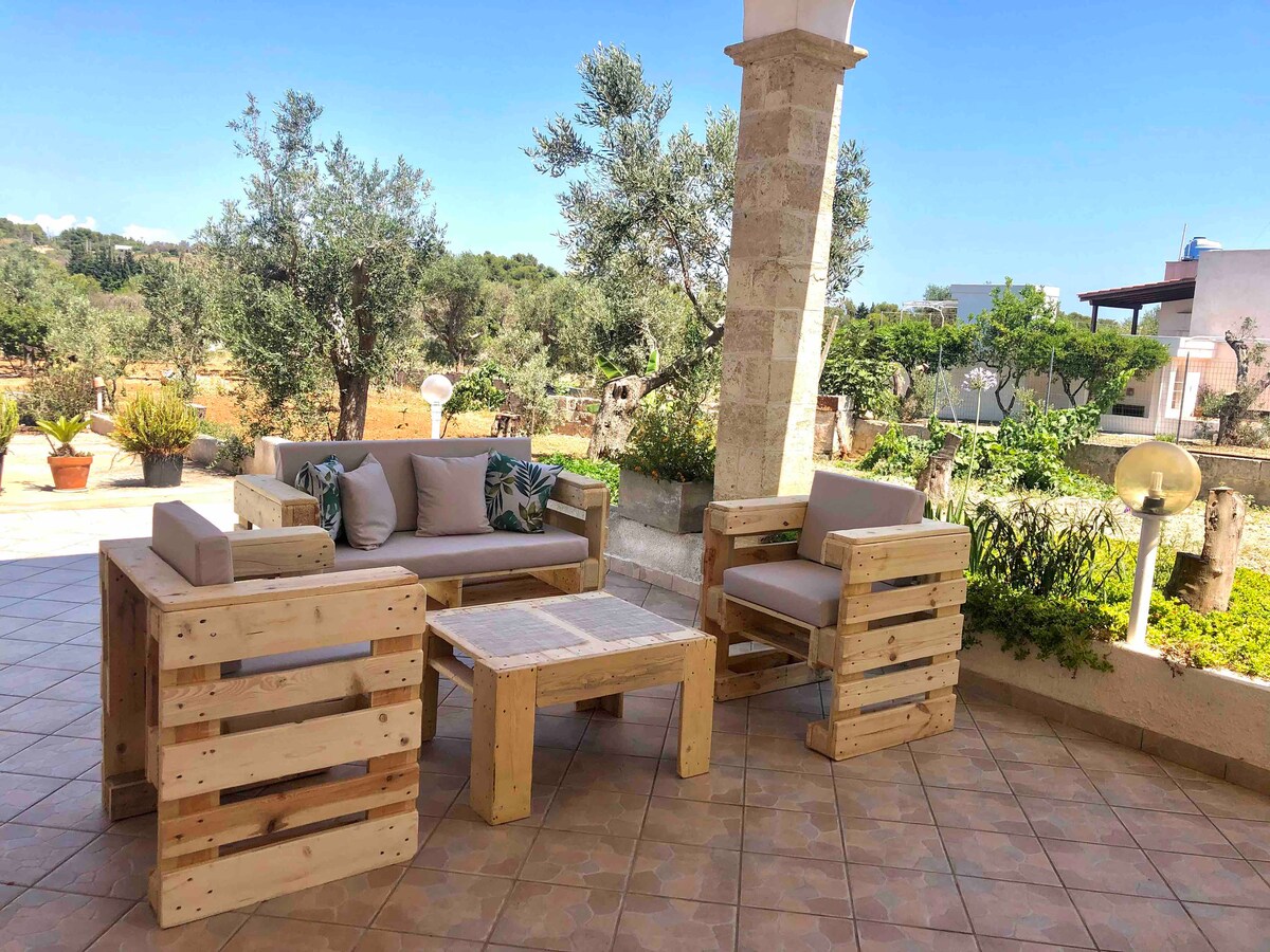An outdoor seating area is showcased, featuring comfortable pallet furniture that includes a sofa and coffee table. The space is surrounded by greenery and offers a tranquil view of the garden and olive trees, enhancing the outdoor experience.