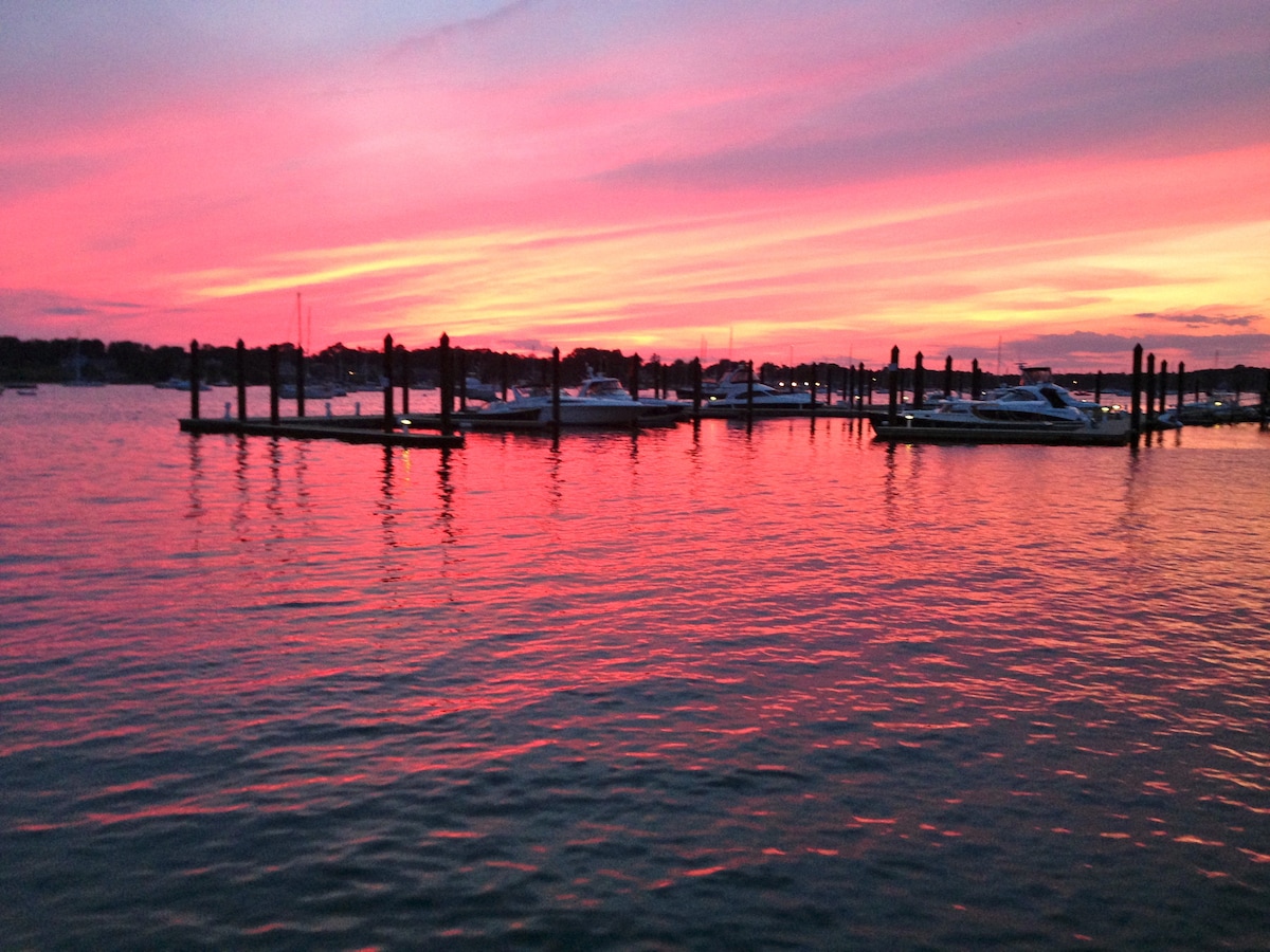 A serene waterfront scene features numerous boats docked amidst reflective waters. A vibrant sunset casts shades of pink and orange across the sky, creating a picturesque backdrop. The silhouettes of docks emerge against the colorful horizon, enhancing the tranquil atmosphere.