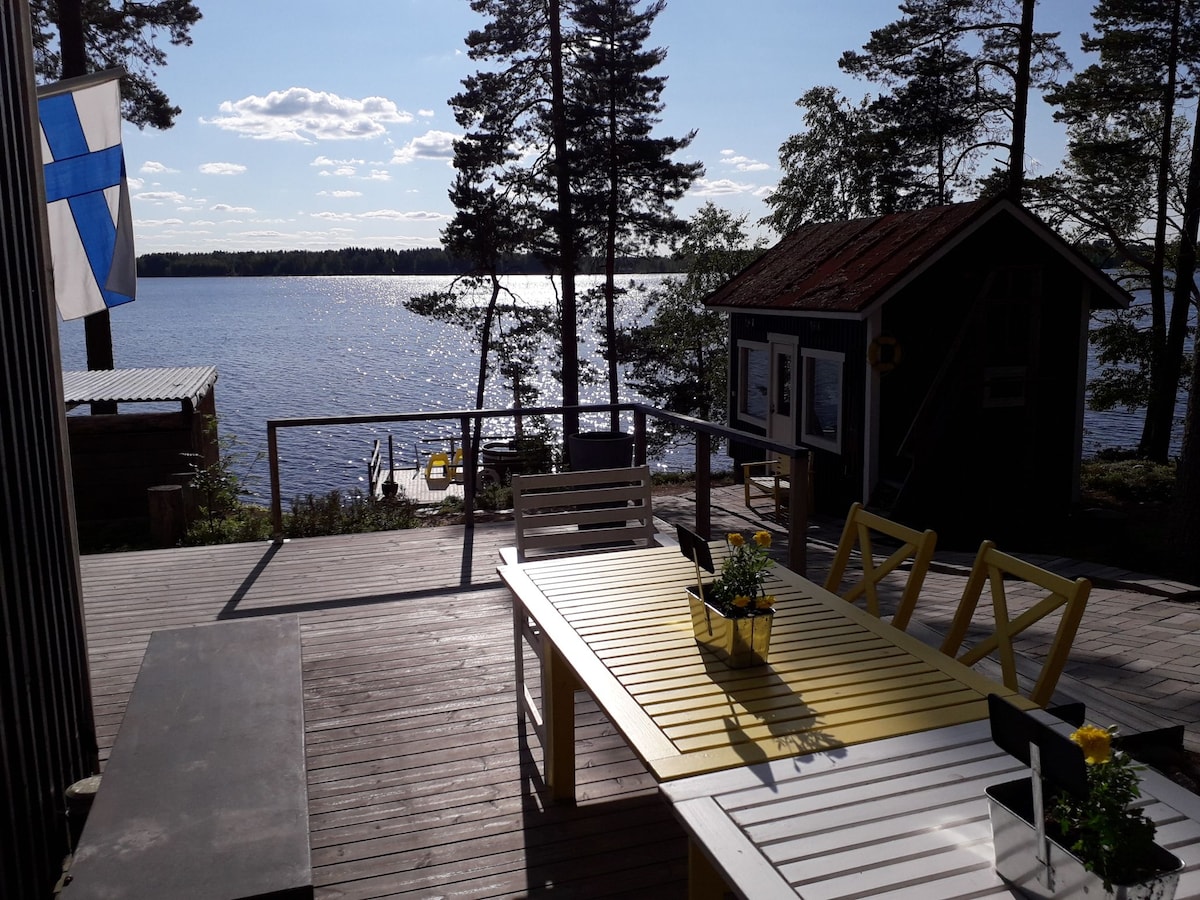 A wooden deck overlooks the lake, featuring a large table with yellow and white chairs, adorned with potted flowers. A small cabin and a Finnish flag are visible, while the water reflects the blue sky and surrounding trees under natural light.