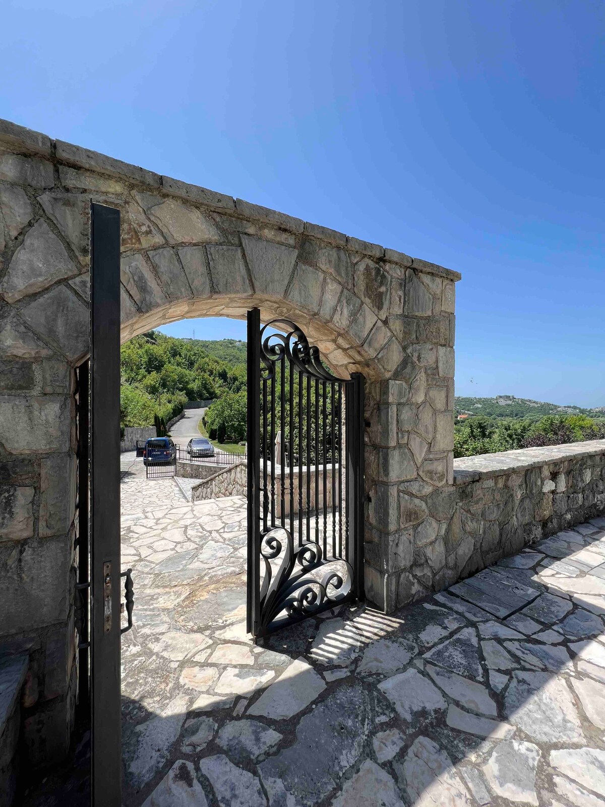 An intricately designed stone archway leads to the villa's entrance, featuring a wrought-iron gate. Sunlight illuminates the surrounding greenery and the path that stretches into the distance, offering a glimpse of the serene landscape beyond.