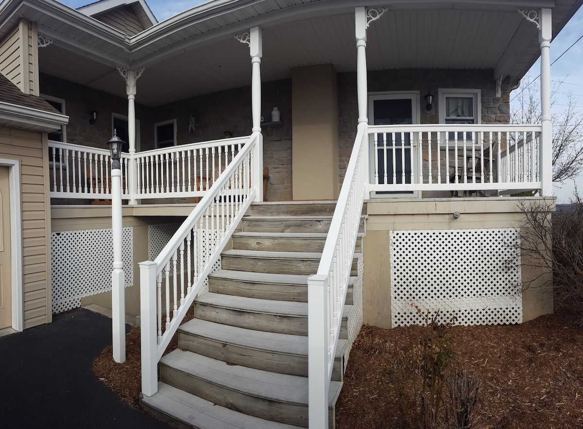 A spacious front porch is featured, complete with a railing and wide steps leading to the entrance. The stone exterior of the house complements the wooden details, providing a welcoming first impression. Surrounding greenery adds a touch of nature to the setting.