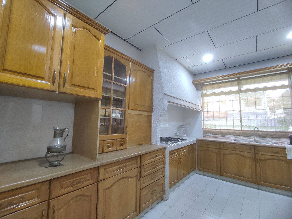 A well-equipped kitchen features wooden cabinetry with a glossy finish, offering ample storage. A large window allows natural light to illuminate the space, while modern appliances, including a gas stove and sink, are visible. The tiled floor adds a clean and functional touch.