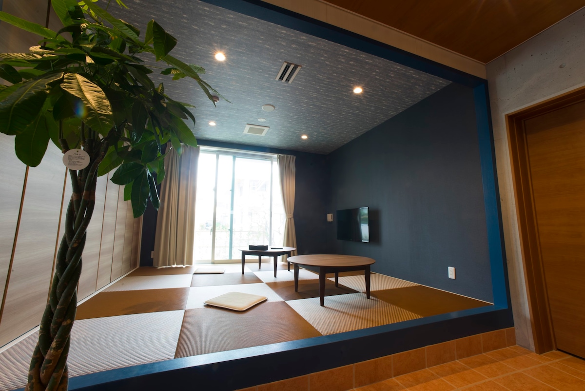 A shared living space is presented with a traditional tatami mat area. A low wooden table is centered in the room, complemented by a floor cushion. Natural light floods in through large sliding glass doors, which are framed by soft curtains.