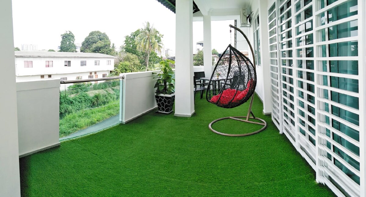 A spacious verandah features an inviting hanging chair with red cushions, surrounded by greenery. A small dining table and chairs are visible, providing a comfortable outdoor setting. The grassy floor offers a soft, natural appearance, creating an enjoyable space for relaxation and gatherings.
