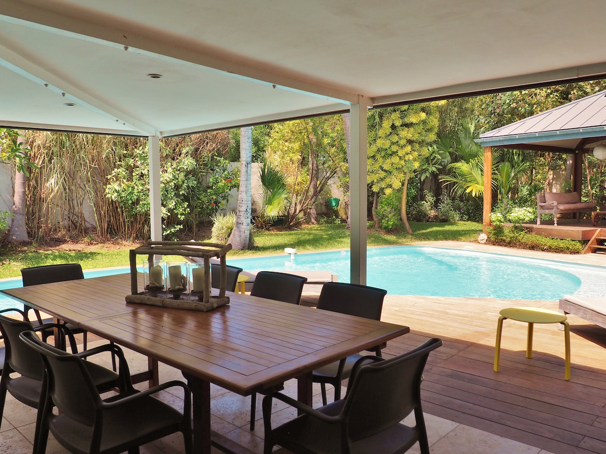 A shaded outdoor dining area features a wooden table surrounded by several chairs, overlooking a tropical garden with lush greenery. A pool is visible in the background, complemented by a gazebo providing additional seating. The serene environment is enhanced by the gentle curvature of the pool.